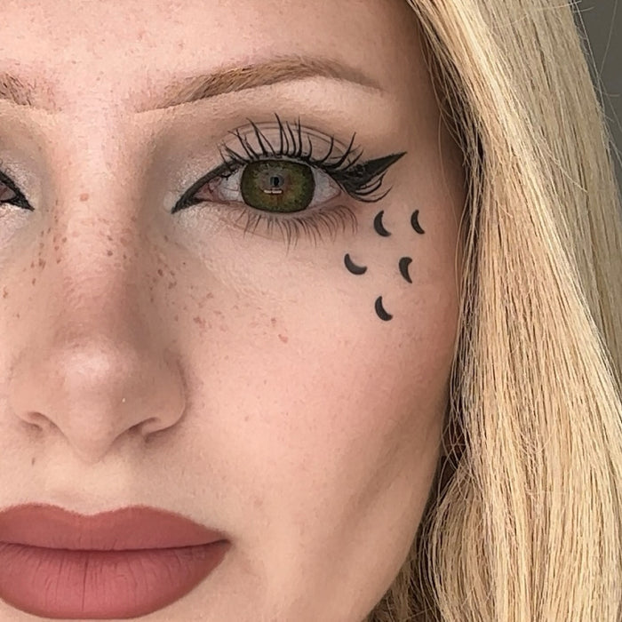 Close-up of a woman's face with a small moon shaped tattoo below her eye.