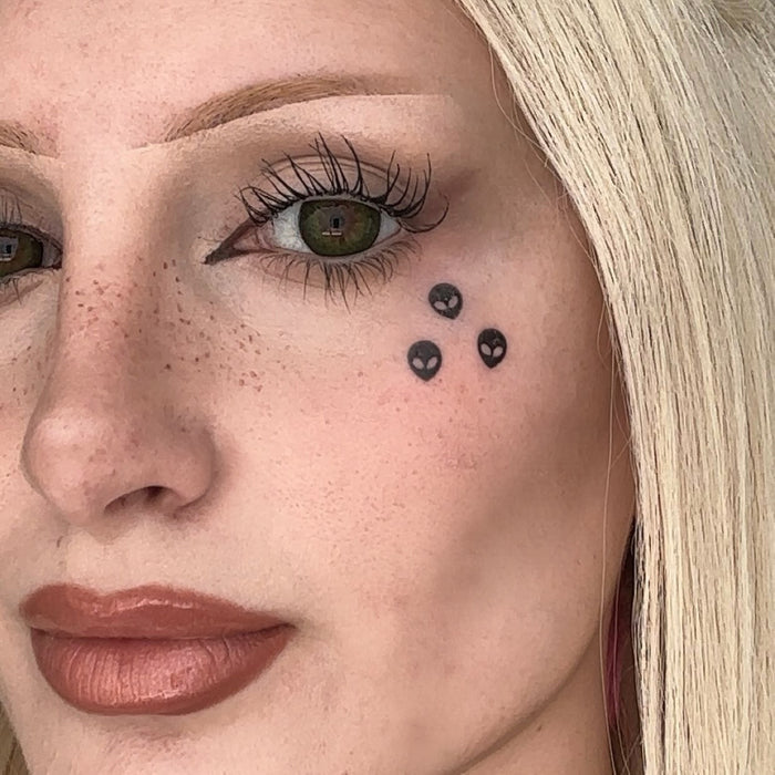 Close-up of a woman's face showing a temporary tattoo of three small black aliens on her cheek.