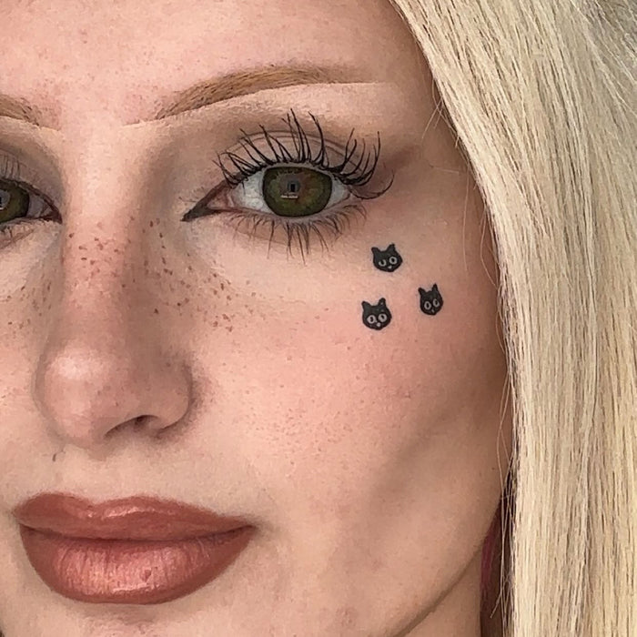 Close-up of a woman's face showing a temporary tattoo of three small black cats on her cheek.