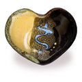 A heart-shaped ceramic bowl with a glossy finish, featuring a split design of warm yellow glaze on one side and a dark brown textured glaze with a blue wavy pattern on the other, set against a plain white background.