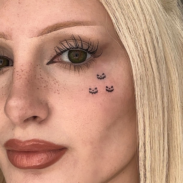 Close-up of a woman's face showing a temporary tattoo of three small black jack o lanterns on her cheek.