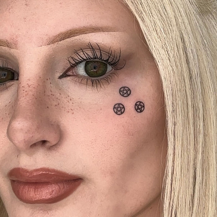 Close-up of a woman's face showing a temporary tattoo of three small black pentagrams on her cheek.